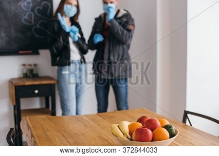 Focus Of Young Couple In Medical Masks And Latex Gloves, And Bowl Of Fresh Fruits On Table
