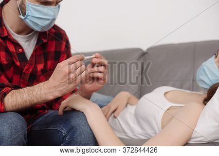View Of Man In Medical Mask Looking At Thermometer Near Sick Girlfriend Lying On Sofa