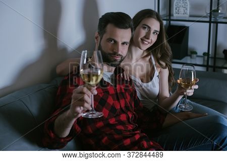 Young Couple Looking At Camera While Sitting On Sofa And Holding Glasses Of White Wine