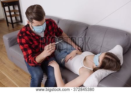 Angle View Of Man In Medical Mask Looking At Thermometer Near Diseased Girlfriend Lying On Sofa