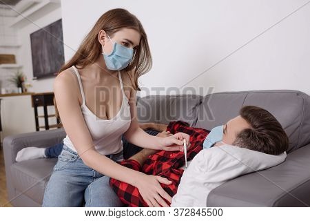 Woman In Medical Mask Holding Thermometer Near Sick Boyfriend Lying On Sofa