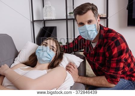 Man In Medical Mask Looking At Camera Near Sick Girlfriend Lying On Sofa