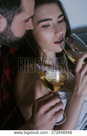 Man Embracing Dreamy Girlfriend While Sitting On Sofa With Glasses Of White Wine