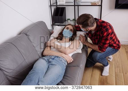 Angle View Of Man Squatting Near Diseased Girlfriend Lying On Sofa In Medical Mask