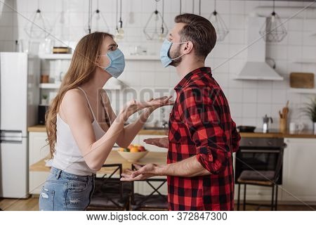 Couple In Medical Masks Quarreling And Gesturing In Kitchen
