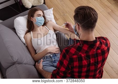 View Of Man Looking At Thermometer Near Diseased Girlfriend In Medical Mask Lying On Sofa