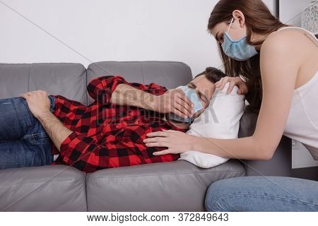 Woman In Medical Mask Squatting Near Ill Boyfriend Lying On Sofa In Medical Mask And Sneezing