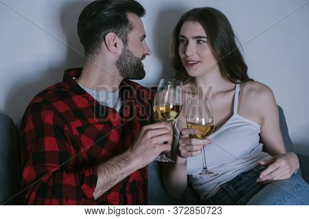 Happy Young Couple Sitting On Sofa, Looking At Each Other While Holding Glasses Of White