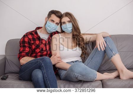 Young Couple In Medical Masks Sitting On Sofa And Looking At Camera