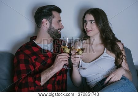 Young Couple Sitting On Sofa, Looking At Each Other And Clinking Glasses Of White Wine