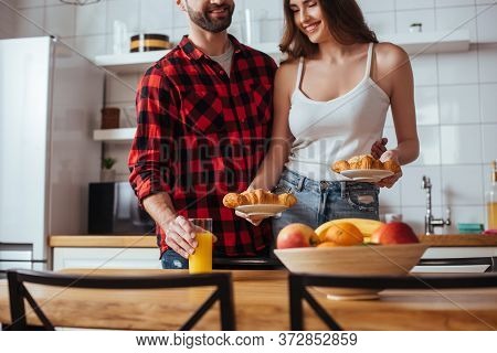 View Of Man Touching Glass Of Orange Juice Near Happy Girlfriend Holding Plates With Delicious Crois