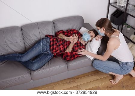 Angle View Of Girl Squatting Near Sick Boyfriend Lying On Sofa In Medical Mask