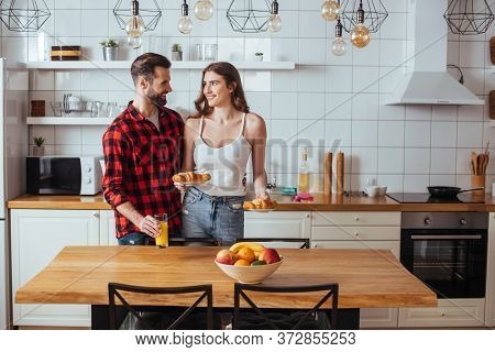 Girl Holding Plates With Croissants While Looking At Smiling Boyfriend Touching Glass Of Orange Juic