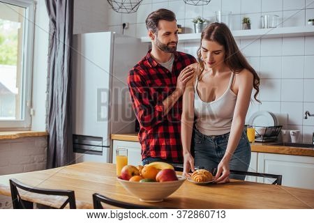 Man Touching Shoulder Of Pretty Girlfriend Holding Plate With Tasty Croissant Near Fresh Fruits