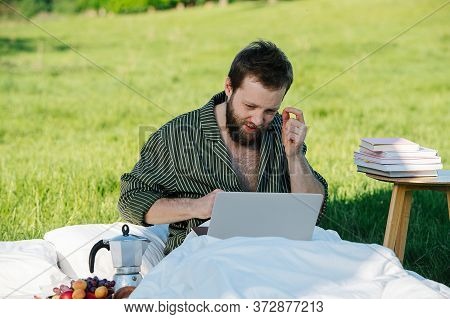 Man Sitting In Bed Outdoors In Grassfield, Working On His Laptop