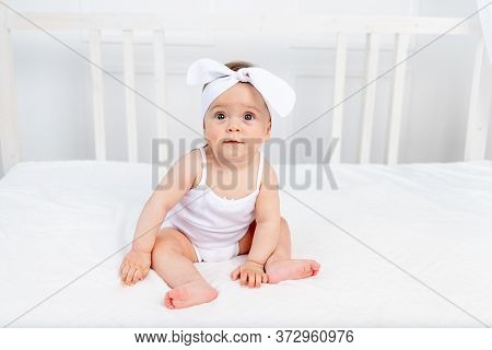 Smiling Baby Girl 8 Months Old Sitting In A Crib In A Children's Room In White Clothes And A Bow And