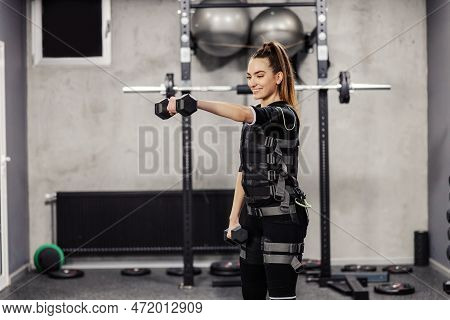 A Sports Girl Wearing An Ems Suit Doing Dumbbell Arm Exercises. A Smile And A Positive Attitude In T