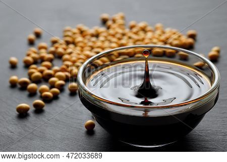 Soy Sauce With Spalsh And Drop In Glass Bowl And Dry Soybeans On Black Stone Background