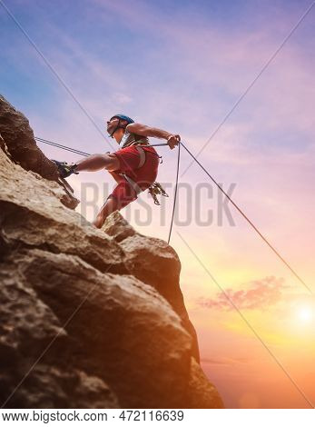 Muscular Climber Man In Protective Helmet Abseiling From Cliff Rock Wall Using Rope Belay Device And