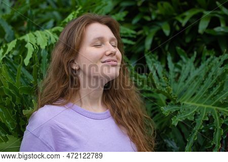 Portrait Of Young Woman Standing On An Ivy, Fern Wall Or In Greenery, Breathing, Smiling And Looking
