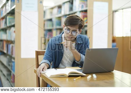Young Male College Student Wearing Eyeglasses And In Casual Cloths Sitting At Desk Studying, Reading