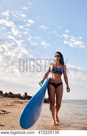 Charming Woman With Dark Hair And Athletic Tanned Body Smiling Carries A Windsurfboard In The Settin