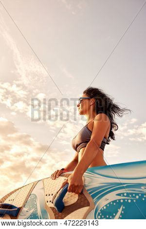 Charming Woman With Dark Hair And Athletic Tanned Body Smiling Carries A Windsurfboard In The Settin