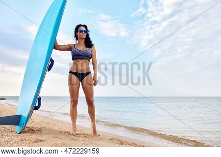 Charming Woman With Dark Hair And Athletic Tanned Body Smiling Carries A Windsurfboard In The Settin