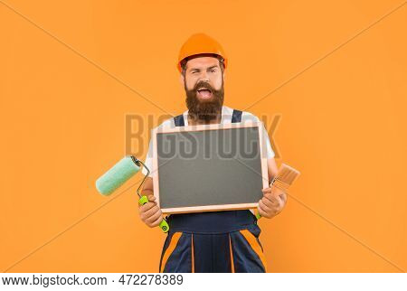 Happy Man Housepainter In Helmet And Work Clothes Hold Paint Roller Brush And Blackboard