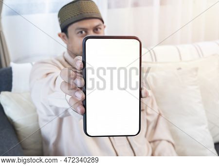 Young Asian Muslim Man In Traditional Clothes Sitting At Sofa In The Living Room And Showing Mobile 