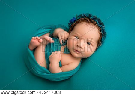 Top View Of A Newborn Baby Girl Lying With Her Eyes Open In A Turquoise Wrap With A Turquoise Headba