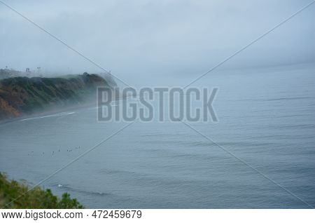 Surfers Ride The Waves In A Fog-shrouded, Misty Sea Cove. Dramatic Cliffs Surround A Secluded Bay, W