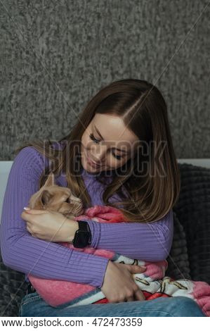 A Young Woman In Her Bedroom Holds Her Pet Cat Wrapped In A Baby Blanket In Her Arms. Love And Care 