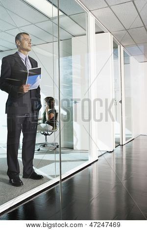 Longitud total del considerado empresario envejecido medio parado contra la pared de cristal en la oficina