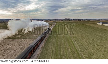 A Behind Aerial View Of A Steam Passenger Train Approaching, Traveling Thru Open Farmlands, Blowing 