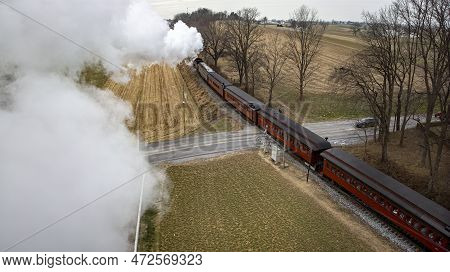 A Behind Aerial View Of A Steam Passenger Train Approaching, Traveling Thru Open Farmlands, Blowing 