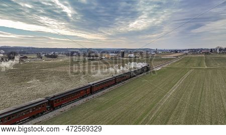 A Behind Aerial View Of A Steam Passenger Train Approaching, Traveling Thru Open Farmlands, Blowing 