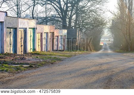 Row of concrete garages with colorful metal doors along a road through urban park with trees. Suburb