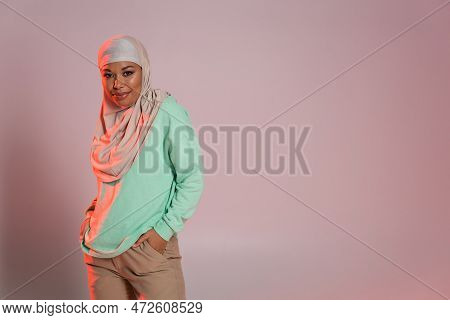 Smiling Multiracial Woman In Hijab And Green Long Sleeve Shirt Posing With Hands In Pockets Of Beige
