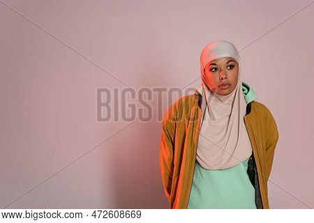 Thoughtful Multiracial Woman In Yellow Jacket And Muslim Hijab Looking Away On Pinkish Grey Backgrou