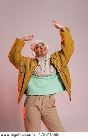 Joyful Multiracial Muslim Woman In Stylish Casual Clothes And Headphones Dancing With Raised Hands O