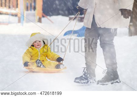 Toddler Baby Rides In The Snow On Icesled, A Winter Playground. Mother Woman Sledding Baby Boy In Ye