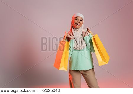 Joyful Multiracial Woman In Trendy Casual Attire And Traditional Hijab Holding Yellow Shopping Bags 