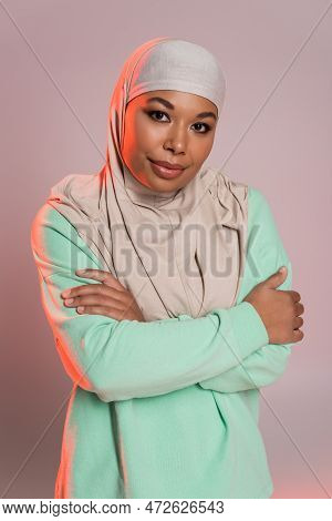 Young Multiracial Woman In Hijab And Green Long Sleeve Shirt Posing With Crossed Arms On Pinkish Gre
