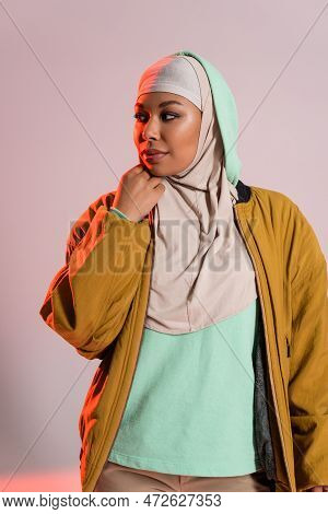 Young And Pretty Multiracial Woman In Yellow Bomber Jacket And Hijab Looking Away On Grey And Pink B