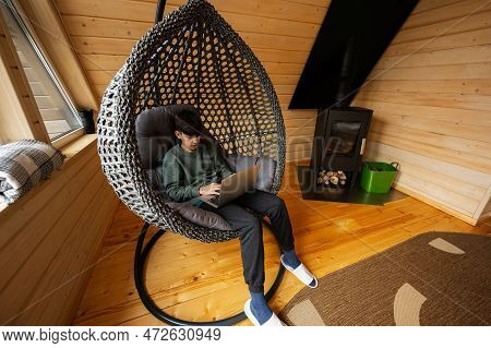 Teenager Boy Sit With Laptop Inside Egg Swing Against Fireplace At Cozy Indoors Heating.