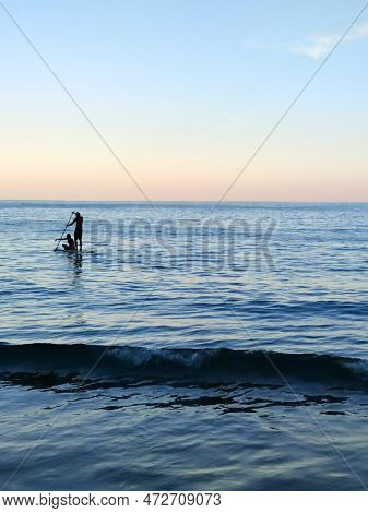 Silhouette Of A Man And A Woman Sailing On A Supboard In The Sea At Sunset.