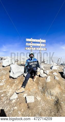 Asian Man Sitting On Top Of A Nearby Mountain Looking At Mount Sindoro. West Java, Indonesia, March