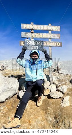 Asian Man Sitting On Top Of A Nearby Mountain Looking At Mount Sindoro. West Java, Indonesia, March