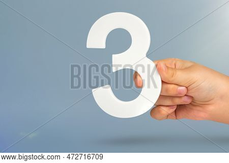 Number Three In Hand. Hand Holding White Number 3 On Blue Background With Copy Space. Concept With N